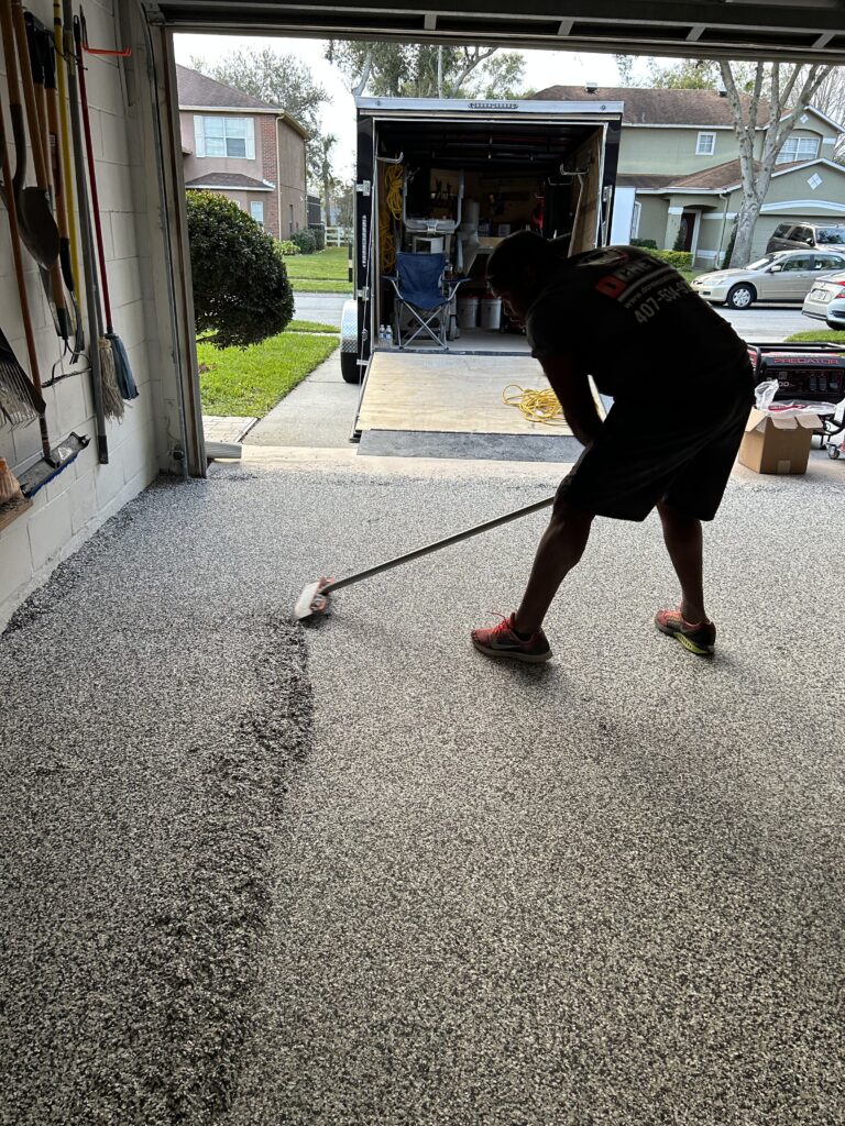 A person applies a textured coating to a garage floor, with tools visible. A trailer and suburban houses are in the background.
