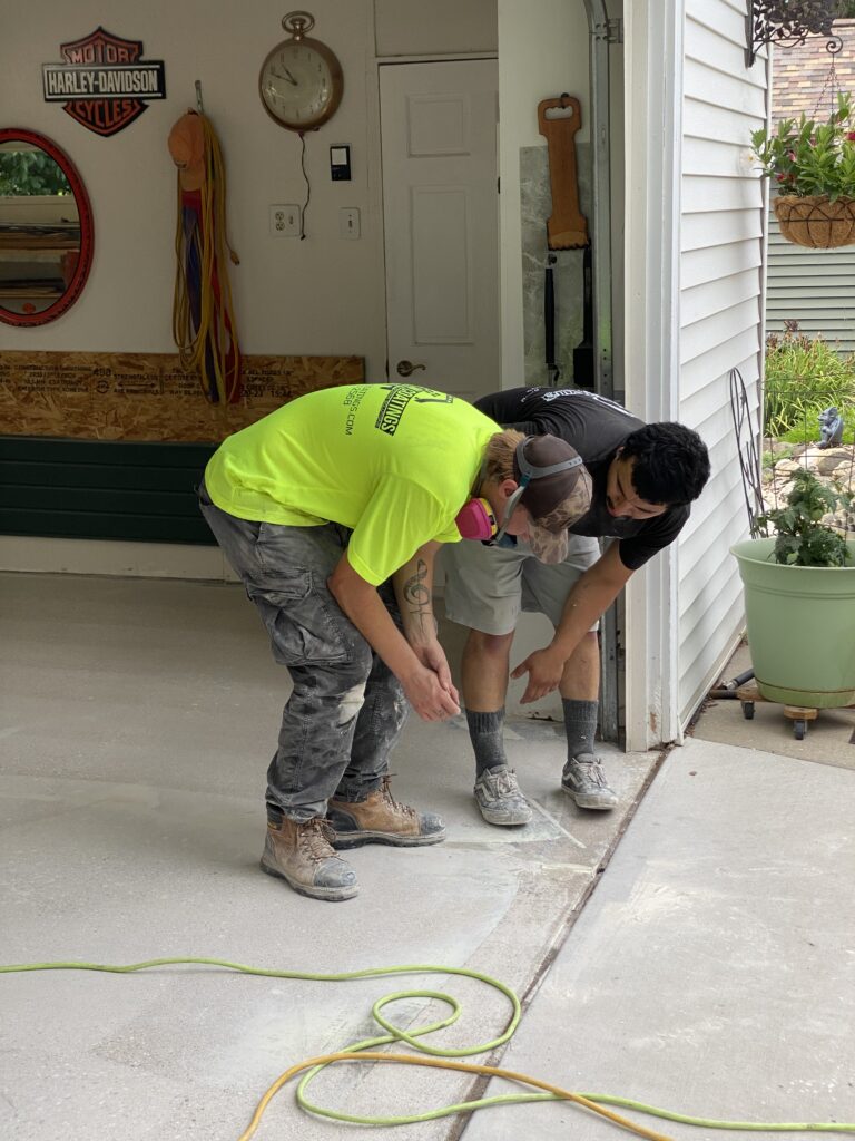 Two people in a garage working on flooring, with a Harley-Davidson sign and plants in the background.