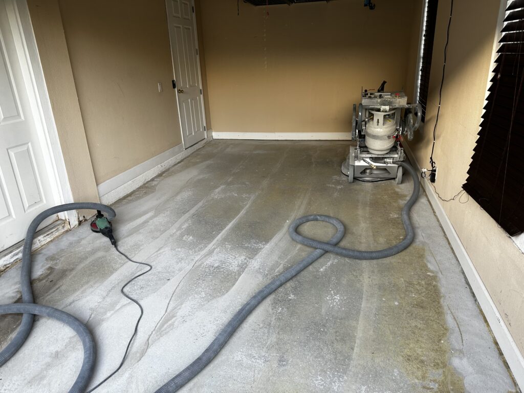 A garage with a concrete floor is being prepared for resurfacing, featuring a floor grinder and a vacuum hose.