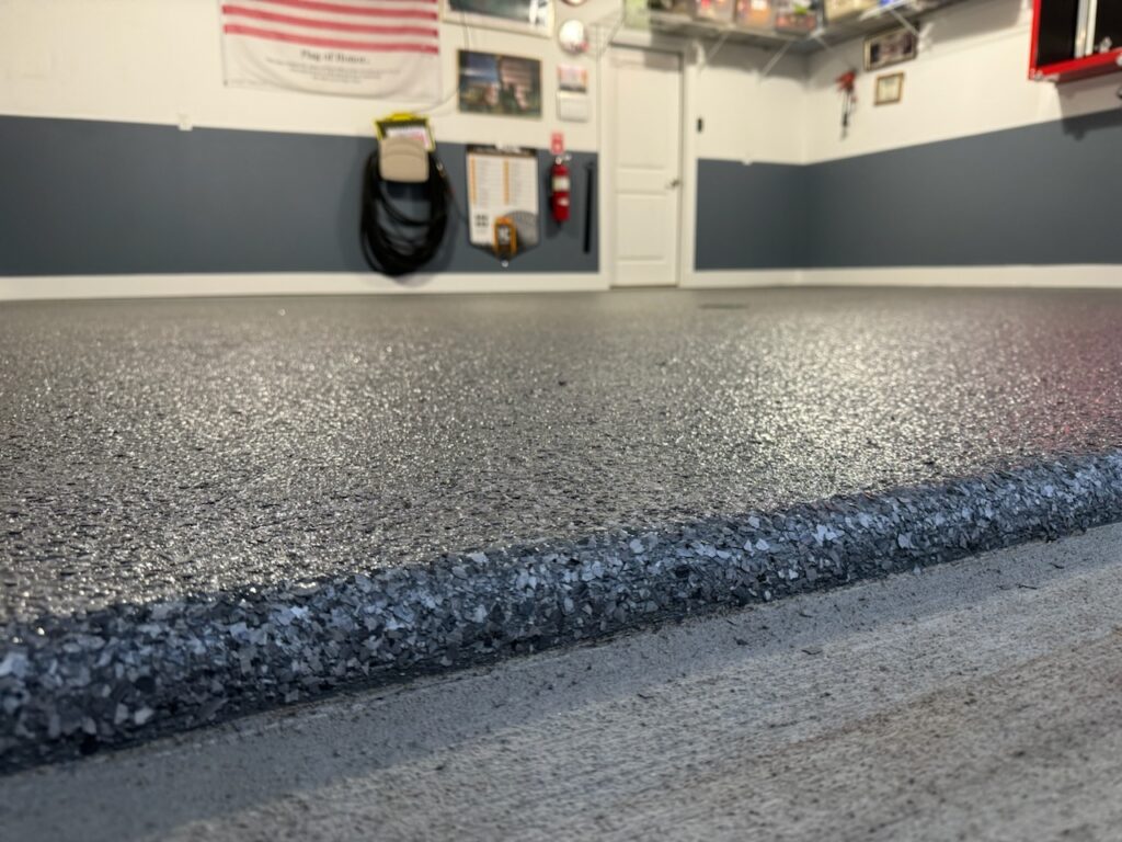 A clean garage interior with a newly coated, speckled epoxy floor. American flag on the wall, storage shelves overhead, door in background.