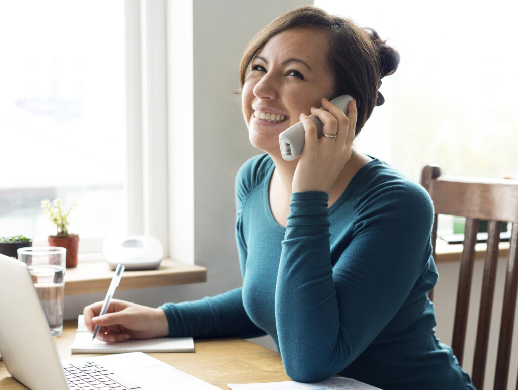 A person with short hair smiles while talking on the phone, writing in a notebook, and using a laptop at a wooden table.