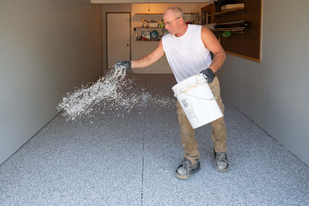 A person in a garage spreads flakes from a bucket onto a freshly coated floor, wearing gloves and work clothes. Shelves in background.
