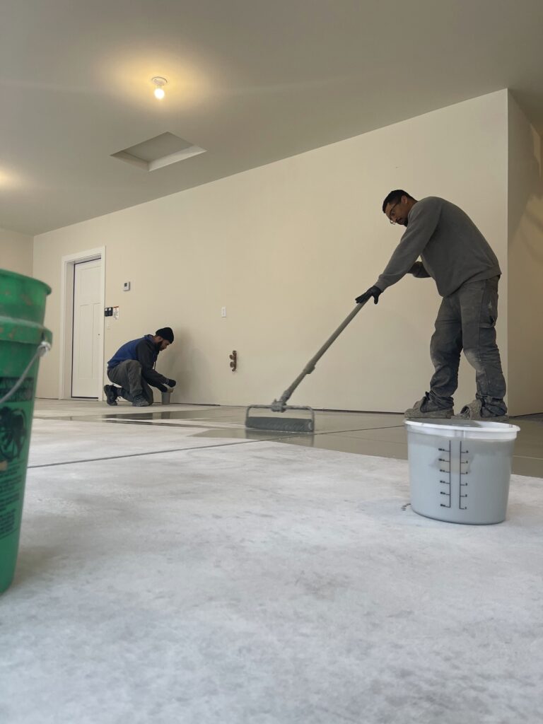 Two people working indoors, painting and applying finishing on a floor. The room is empty, with buckets and minimal lighting.