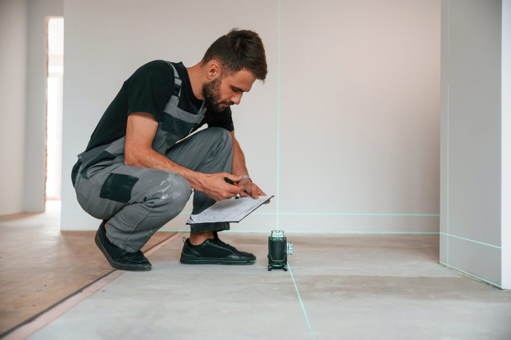 A person in work attire uses a clipboard and laser level to inspect a room with unfinished floors and walls.