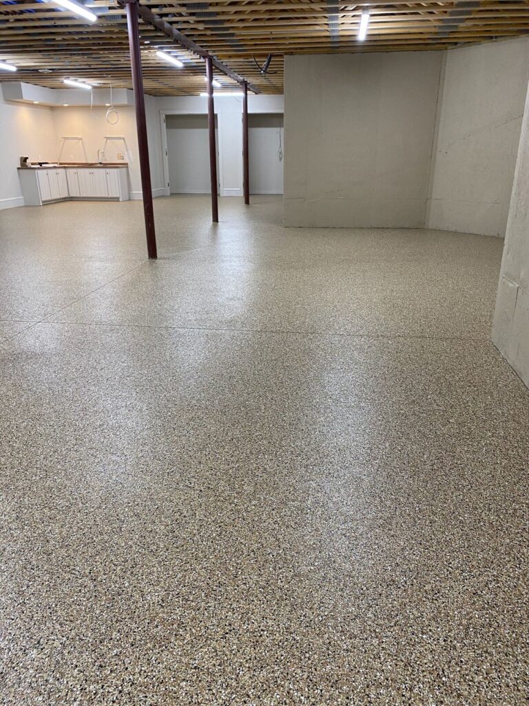 Unfinished basement with exposed ceiling joists, central support columns, and a small kitchenette area. Bright overhead lighting illuminates speckled flooring.