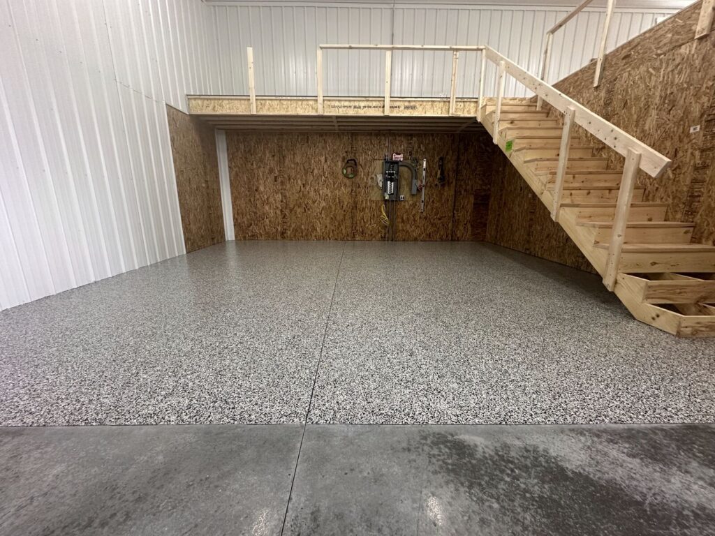A spacious, unfinished room features a wooden staircase leading to a loft. The walls are white and brown, with a speckled floor.